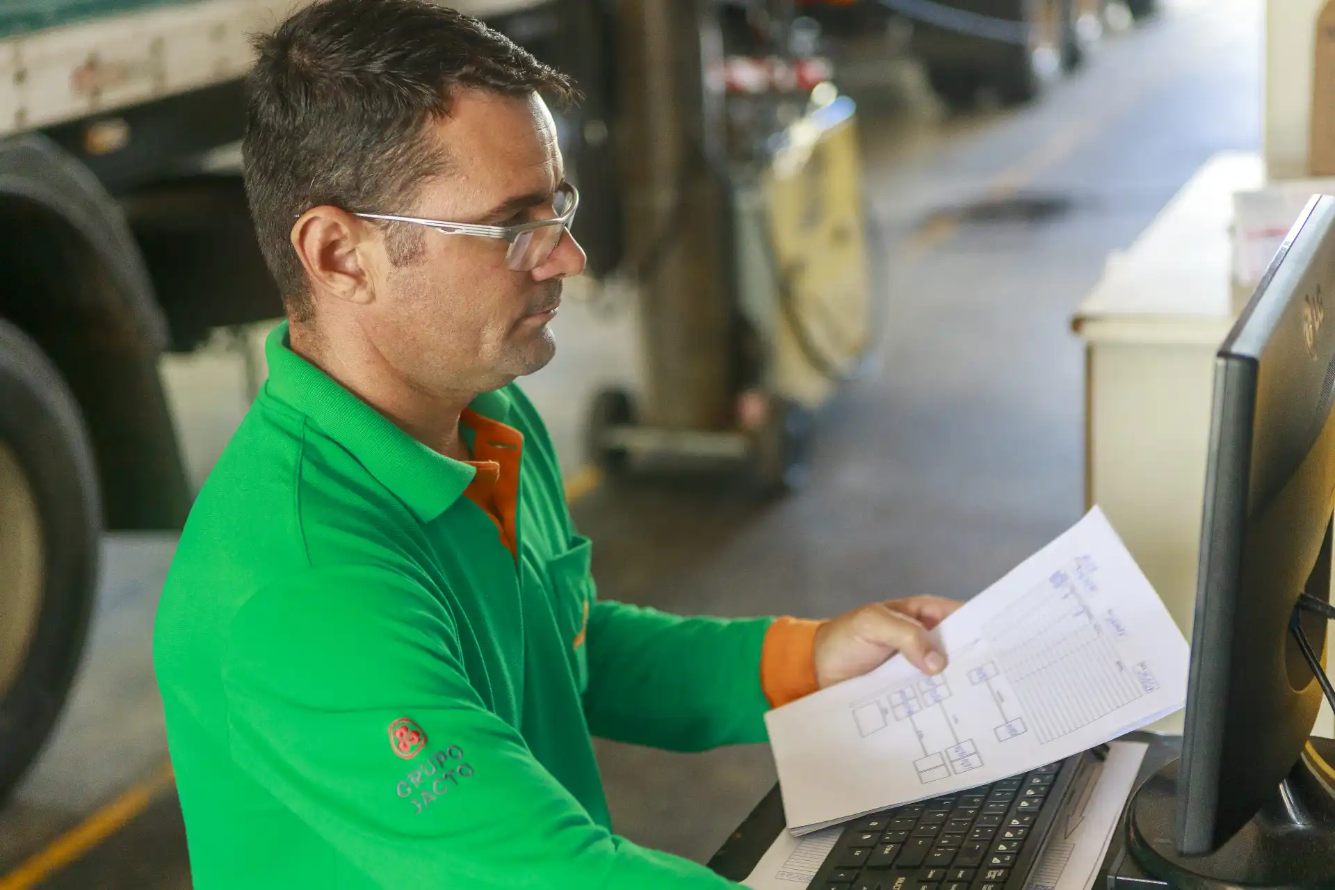 Homem exerce função de monitoramento ou controle da cadeia logística em ambiente industrial, usando equipamento de computador, vestido com uniforme verde com detalhes laranja.